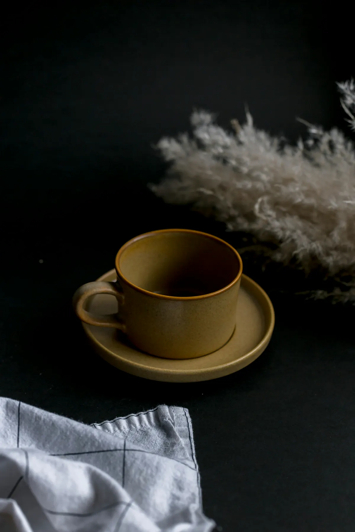 A mustard ceramic cup on its saucer in low light, a folded white cloth and dry grasses beside it